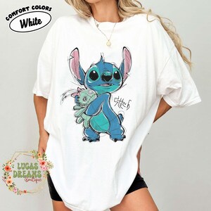 May include: A white short-sleeve t-shirt featuring a blue Stitch character illustration holding a small green doll. The graphic includes the text Stitch in a stylized font. The shirt has a Comfort Colors label printed on the upper left side.