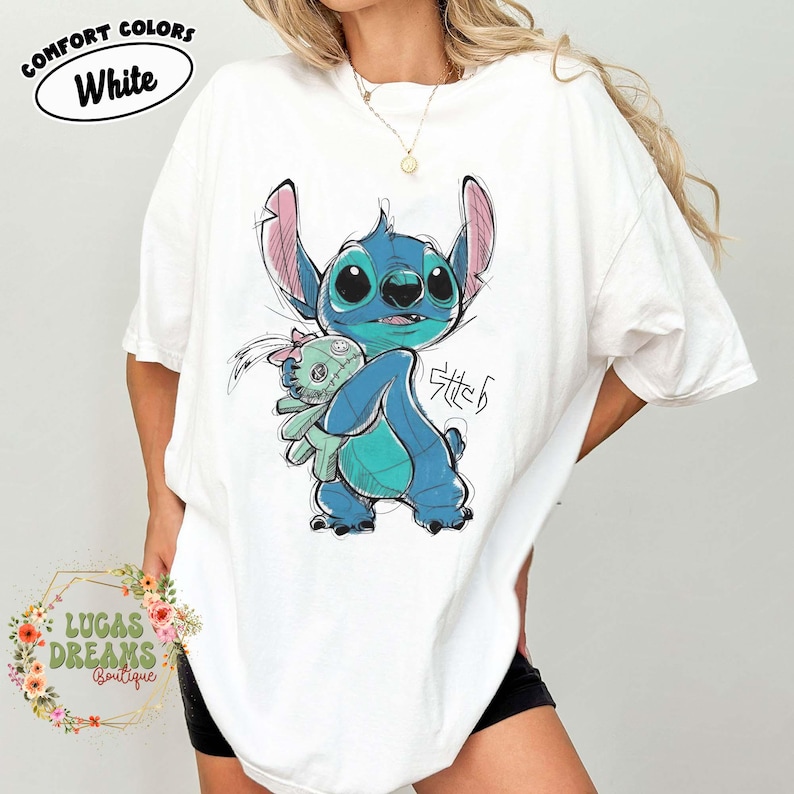 May include: A white short-sleeve t-shirt featuring a blue Stitch character illustration holding a small green doll. The graphic includes the text Stitch in a stylized font. The shirt has a Comfort Colors label printed on the upper left side.