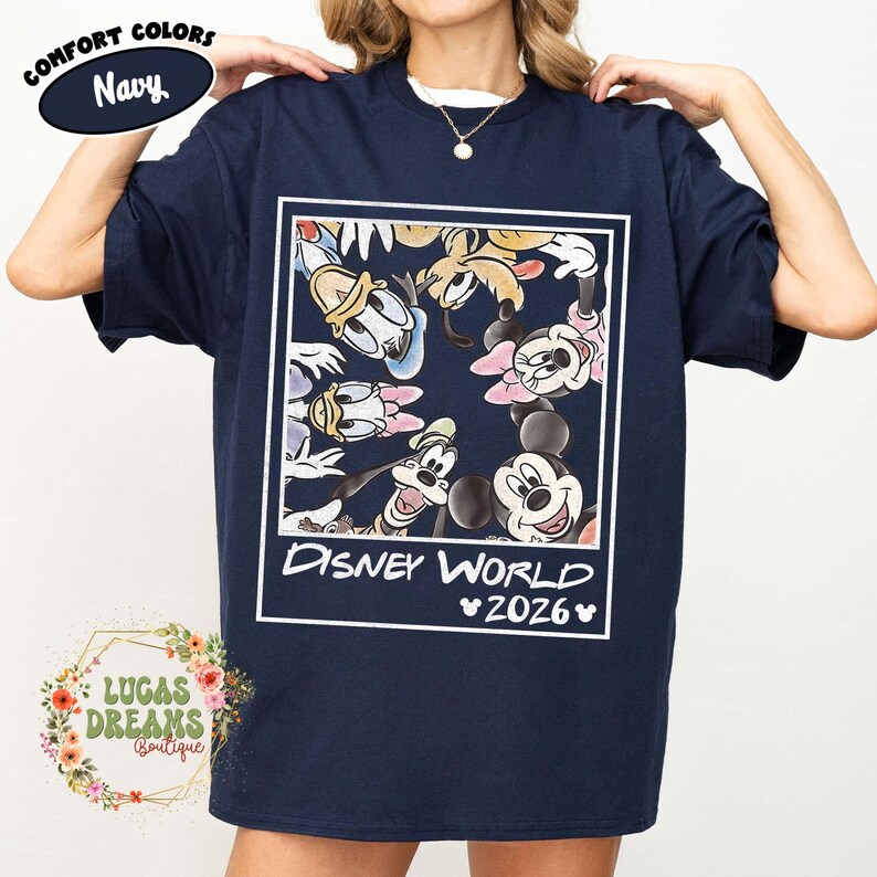 Op de afbeelding: Marineblauwe t-shirt met een omlijnde afbeelding van Disney-personages zoals Mickey Mouse, Minnie Mouse, Donald Duck en Goofy. De tekst "Disney World 2026" staat eronder. Het shirt heeft de tekst "Comfort Colors Navy" linksboven.