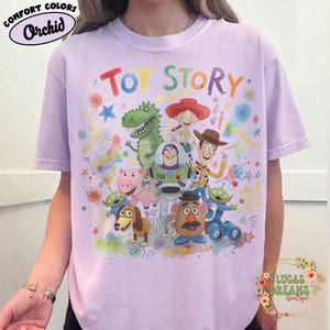 Pu&ograve; includere: T-shirt lilla chiaro con la scritta "TOY STORY" e illustrazioni di personaggi. La maglietta presenta anche le parole "COMFORT COLORS" e "Orchid".