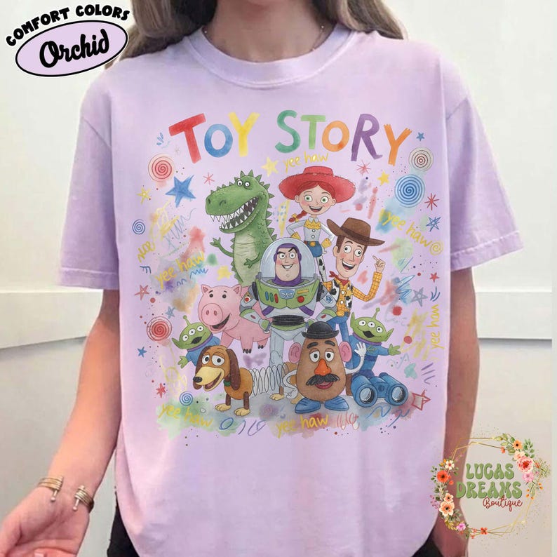 Pu&ograve; includere: T-shirt lilla chiaro con la scritta "TOY STORY" e illustrazioni di personaggi. La maglietta presenta anche le parole "COMFORT COLORS" e "Orchid".