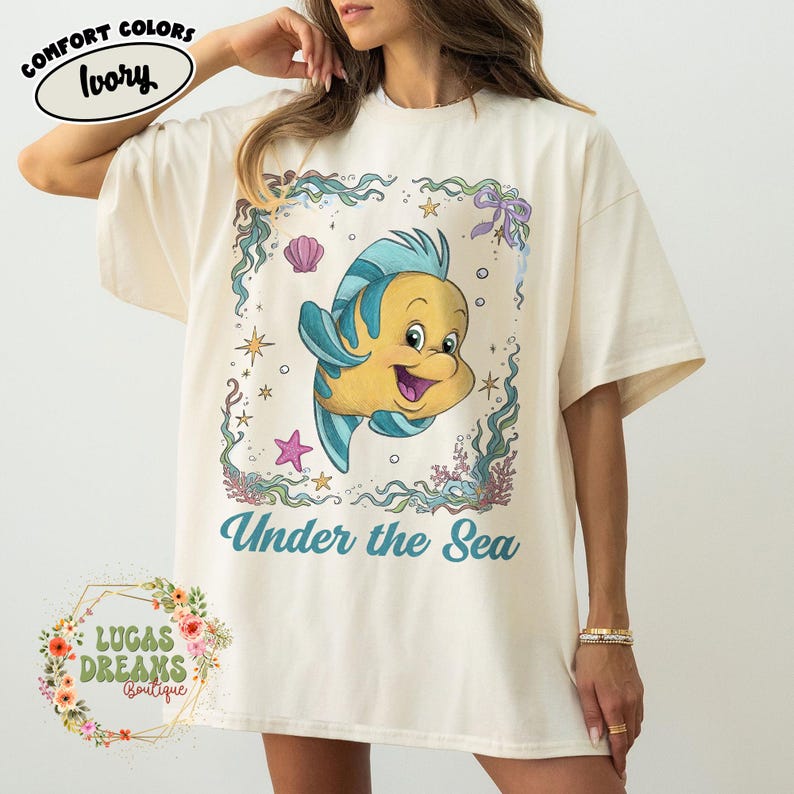 May include: Ivory-colored t-shirt featuring a cartoon image of a yellow and blue fish with the text "Under the Sea." The design is framed with aquatic elements like seaweed, shells, and stars. The shirt is from Lucas Dreams Boutique.