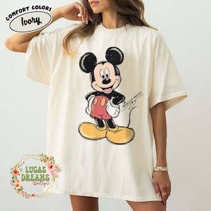 May include: An ivory short-sleeve t-shirt featuring a graphic of Mickey Mouse with black ears, red shorts, and yellow shoes. The design includes the text MICKEY MOUSE in a script font. A label in the top left corner reads COMFORT COLORS IVORY.