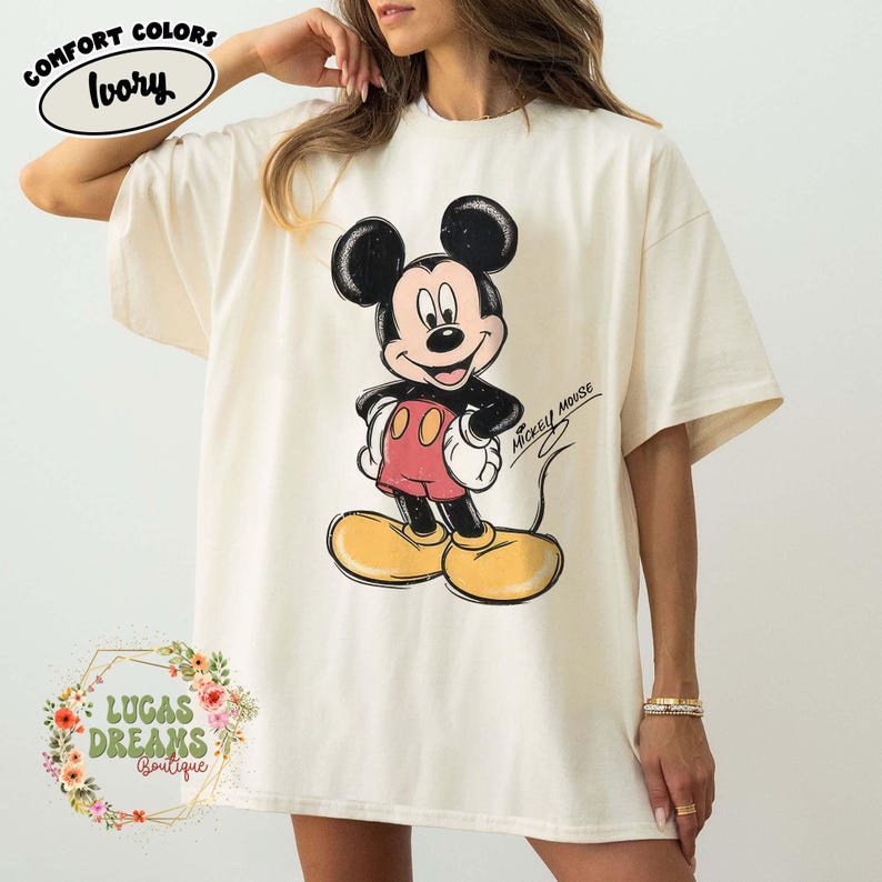 May include: An ivory short-sleeve t-shirt featuring a graphic of Mickey Mouse with black ears, red shorts, and yellow shoes. The design includes the text MICKEY MOUSE in a script font. A label in the top left corner reads COMFORT COLORS IVORY.