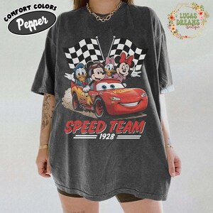 Op de afbeelding: Donkergrijs T-shirt met een afbeelding van Disney-personages in een rode raceauto met geblokte vlaggen. De tekst "SPEED TEAM 1928" staat onder de auto. Op de shirt staat linksboven "COMFORT COLORS Pepper".