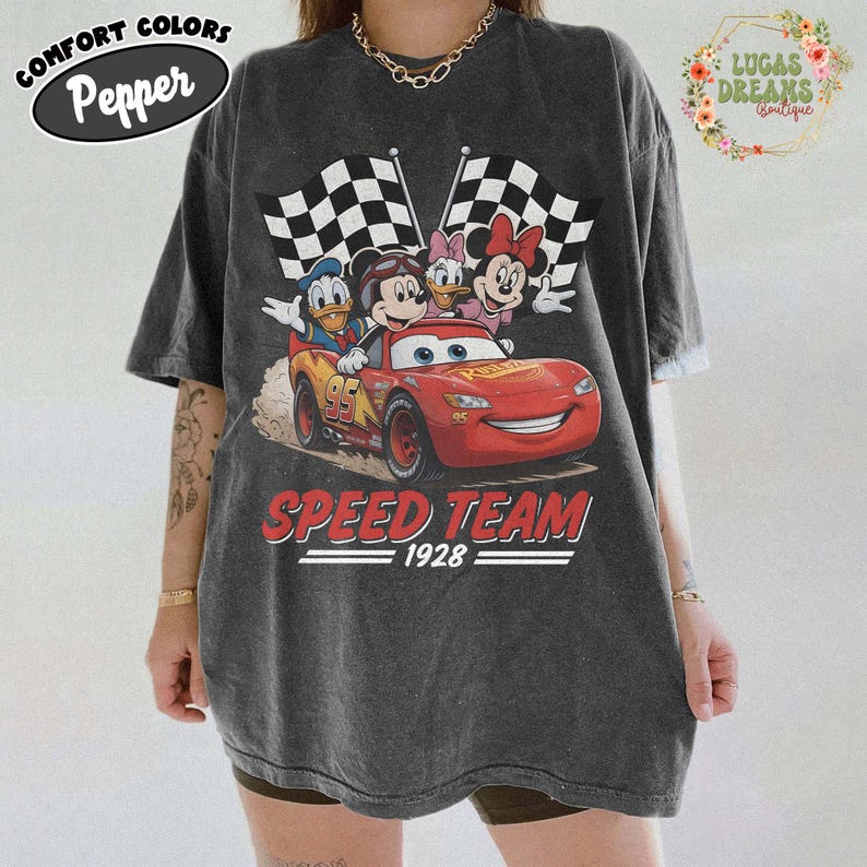 Op de afbeelding: Donkergrijs T-shirt met een afbeelding van Disney-personages in een rode raceauto met geblokte vlaggen. De tekst "SPEED TEAM 1928" staat onder de auto. Op de shirt staat linksboven "COMFORT COLORS Pepper".