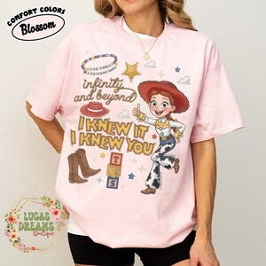 May include: A light pink t-shirt featuring a graphic of a cowgirl character, a red hat, brown boots, and alphabet blocks. The text reads infinity and beyond, I KNEW IT I KNEW YOU, and YOU'VE GOT A FRIEND IN ME. The shirt also includes small star and cloud icons.