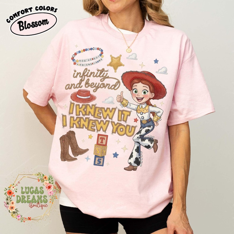 May include: A light pink t-shirt featuring a graphic of a cowgirl character, a red hat, brown boots, and alphabet blocks. The text reads infinity and beyond, I KNEW IT I KNEW YOU, and YOU'VE GOT A FRIEND IN ME. The shirt also includes small star and cloud icons.