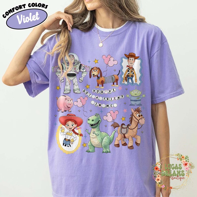 May include: Violet Comfort Colors t-shirt featuring various Toy Story characters. The design includes Buzz Lightyear, Woody, Jessie, and others, with the phrase "YOU'VE GOT A FRIEND IN ME". The shirt has a relaxed fit.
