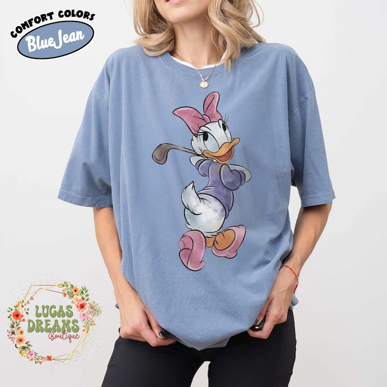 May include: Blue t-shirt with a Daisy Duck graphic. The cartoon character is wearing a purple outfit and pink bow and shoes, holding a golf club. The shirt has the text "COMFORT COLORS Blue Jean" and "LUCAS DREAMS Boutique".