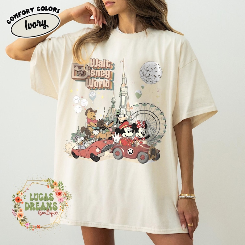 May include: Ivory-colored t-shirt featuring a vintage-style Walt Disney World graphic. The design includes Mickey and Minnie Mouse, Winnie the Pooh, and other characters in red cars, with a Ferris wheel and castle in the background. The shirt has the text "Comfort Colors" and "Ivory".