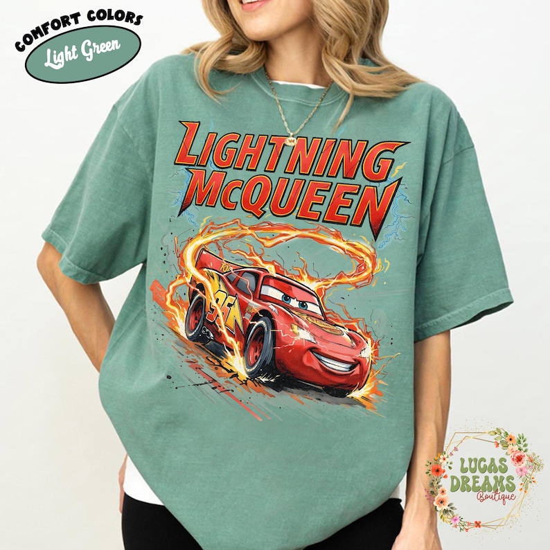 May include: Light green t-shirt featuring a graphic of Lightning McQueen from the Cars movie. The design includes the text "LIGHTNING McQUEEN" in red and yellow, with the character in motion. The shirt also has the text "COMFORT COLORS" and "Light Green".