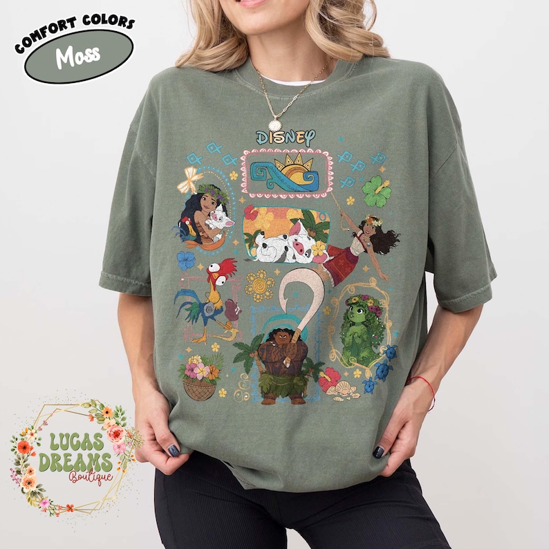 May include: A moss green t-shirt with Disney's Moana characters. The shirt has a crew neck and short sleeves. The word "Disney" is printed across the chest. The shirt also has the text "Comfort Colors" and "Lucas Dreams Boutique".