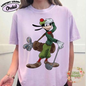May include: A light purple t-shirt featuring a cartoon character dressed as a golfer. The character is wearing a green outfit, a red bow tie, and a golf hat. The shirt has the words "Comfort Colors" and "Orchid" printed on it.