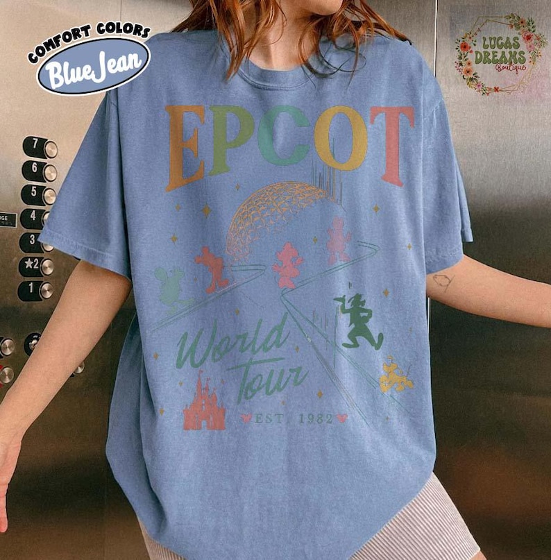 Puede incluir: Camiseta azul claro de manga corta con el texto "EPCOT World Tour EST 1982" en letras y gr&aacute;ficos coloridos. La camiseta presenta un gr&aacute;fico de la bola EPCOT y personajes de dibujos animados.