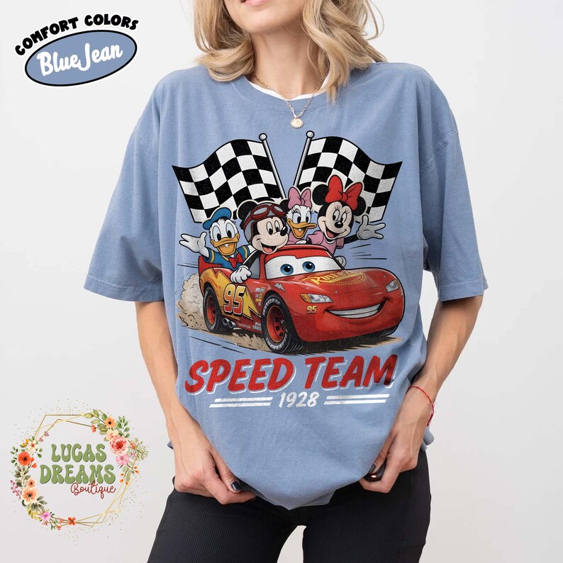 Op de afbeelding: Blauwkleurig T-shirt met korte mouwen en een afbeelding van Disney-personages in een rode raceauto. De auto heeft het nummer 95. De woorden "SPEED TEAM 1928" staan eronder gedrukt. Het shirt heeft de woorden "COMFORT COLORS Blue Jean" erop gedrukt.