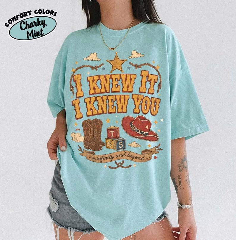 May include: A light mint green short-sleeve t-shirt featuring a graphic print with the text I KNEW IT I KNEW YOU and infinity and beyond. The design includes a brown cowboy boot, a red cowboy hat, a star, and alphabet blocks labelled T, S, and 5.