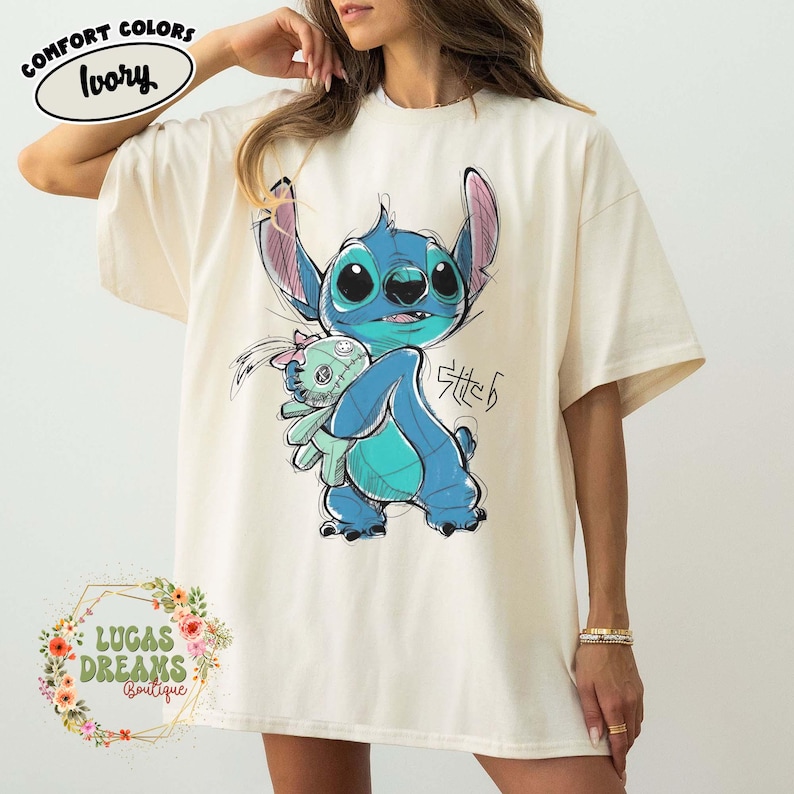 May include: An ivory-colored t-shirt featuring a graphic illustration of the blue character Stitch holding a small doll. The design includes the text Stitch in a stylized script. A Comfort Colors Ivory label is visible in the upper left corner.