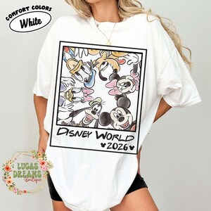 Op de afbeelding: Wit T-shirt met een ingelijste afbeelding van Disney-personages, waaronder Mickey en Minnie Mouse, Donald Duck en Goofy. De tekst "Disney World 2026" staat eronder. Het shirt heeft de woorden "Comfort Colors" en "White" in een cirkel.