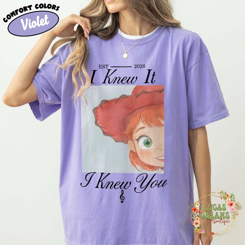 May include: A violet short-sleeved t-shirt featuring a graphic print of a character with red hair and a red hat. The text reads "EST 2026 I Knew It" above the graphic and "I Knew You" below it, with a small musical note symbol between the words.