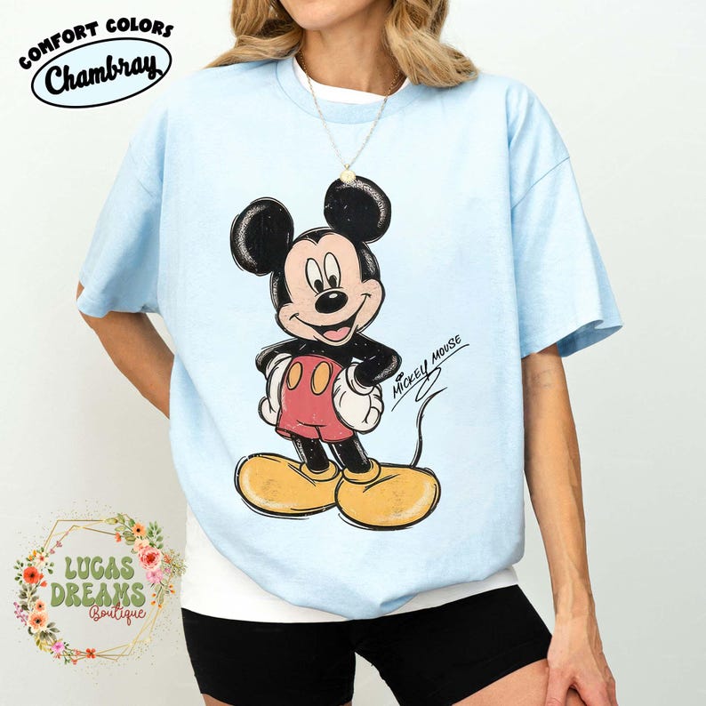 May include: A light blue short-sleeve t-shirt featuring a graphic of Mickey Mouse with black ears, red shorts, and yellow shoes. The text "MICKEY MOUSE" is printed in black script next to the character. The shirt has a crew neckline and a loose fit.