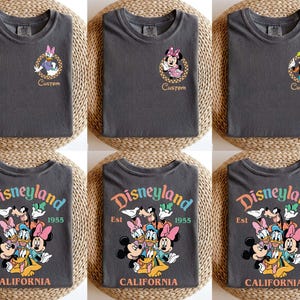 Puede incluir: Camisetas gris oscuro con personajes de Disney. Algunas camisetas tienen retratos de personajes con la palabra "Custom". Otras muestran "Disneyland Est 1955 California" con personajes de Disney. Las camisetas est&aacute;n dobladas sobre una superficie tejida.