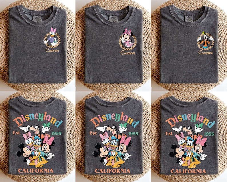 Puede incluir: Camisetas gris oscuro con personajes de Disney. Algunas camisetas tienen retratos de personajes con la palabra "Custom". Otras muestran "Disneyland Est 1955 California" con personajes de Disney. Las camisetas est&aacute;n dobladas sobre una superficie tejida.