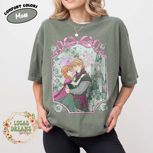 Puede incluir: Camiseta verde musgo con una ilustraci&oacute;n enmarcada de dos personajes animados abraz&aacute;ndose, con un castillo de fondo. El marco incluye un lazo rosa y rosas. El texto "COMFORT COLORS Moss" est&aacute; en la parte superior y "LUCAS DREAMS Boutique" en la parte inferior.