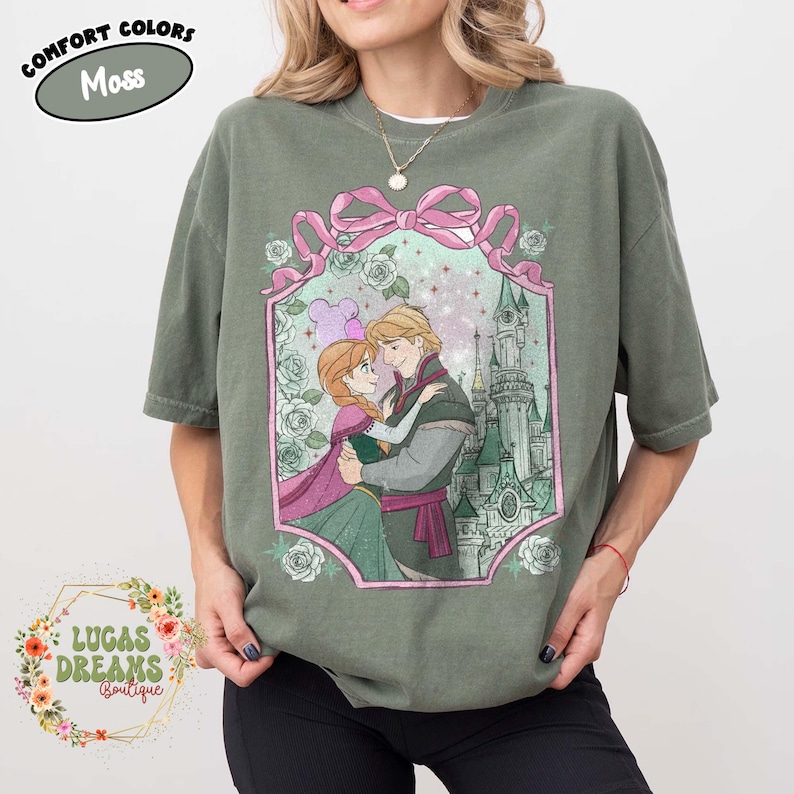 Puede incluir: Camiseta verde musgo con una ilustraci&oacute;n enmarcada de dos personajes animados abraz&aacute;ndose, con un castillo de fondo. El marco incluye un lazo rosa y rosas. El texto "COMFORT COLORS Moss" est&aacute; en la parte superior y "LUCAS DREAMS Boutique" en la parte inferior.