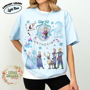 Könnte beinhalten: Hellblaues T-Shirt mit Figuren aus dem Film Frozen. Die Grafik zeigt Elsa, Anna, Olaf und andere Charaktere, mit dem Schriftzug "Let It Go". Das Shirt ist mit "Comfort Colors" und "Light Blue" gekennzeichnet.