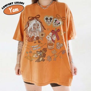 May include: Orange t-shirt with a graphic featuring a castle, Mickey and Minnie Mouse, Chip and Dale, and fall-themed elements. The shirt has the text "Comfort Colors" and "Yam" in an oval. The design includes pumpkins, acorns, and leaves.