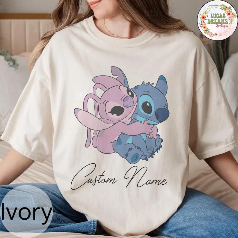 May include: Ivory-colored t-shirt featuring a cartoon image of two characters embracing. One character is blue, the other is pink. The words "Custom Name" are written in cursive below the image. The word "Ivory" is printed at the bottom left.