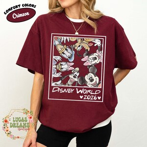 Op de afbeelding: Karmizijnrood T-shirt met een witte ronde hals en een Disney World 2026-afbeelding. De afbeelding bevat Mickey Mouse, Minnie Mouse, Donald Duck, Goofy en andere stripfiguren. Het shirt heeft korte mouwen en een relaxte pasvorm.