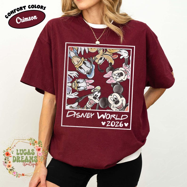 Op de afbeelding: Karmizijnrood T-shirt met een witte ronde hals en een Disney World 2026-afbeelding. De afbeelding bevat Mickey Mouse, Minnie Mouse, Donald Duck, Goofy en andere stripfiguren. Het shirt heeft korte mouwen en een relaxte pasvorm.