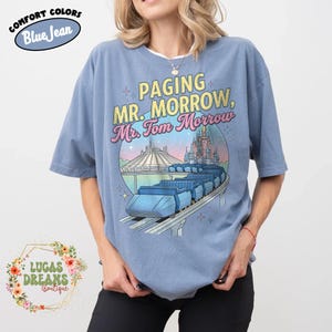 May include: Pale blue t-shirt featuring the text "Paging Mr. Morrow, Mr. Tom Morrow" and a graphic of a futuristic train, Space Mountain, and a castle. The shirt is a comfort colour and has the words "Comfort Colors" and "Blue Jean" on it.