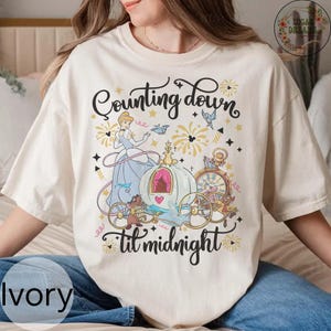 Può includere: T-shirt color avorio con grafica a tema Cenerentola. Il design include Cenerentola, una carrozza e la scritta "Counting down 'til midnight". La grafica è nei toni del blu, oro e marrone.