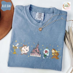 Puede incluir: Camiseta azul vaquero Comfort Colors con un gráfico de personajes y un castillo de cuento de hadas. La camiseta tiene cuello redondo y mangas cortas. Un collar dorado con un colgante está drapeado sobre la camiseta.