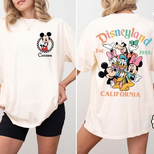 Puede incluir: Camiseta color marfil con Mickey Mouse en un c&iacute;rculo a cuadros y la palabra "Custom". La parte trasera muestra la palabra "Disneyland" en colores arco&iacute;ris, personajes de Disney y el texto "Est 1955 California".