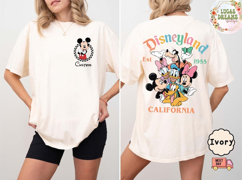 Puede incluir: Camiseta color marfil con Mickey Mouse en un c&iacute;rculo a cuadros y la palabra "Custom". La parte trasera muestra la palabra "Disneyland" en colores arco&iacute;ris, personajes de Disney y el texto "Est 1955 California".