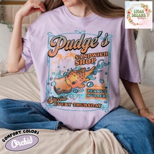 Pu&ograve; includere: T-shirt color orchidea con grafica retr&ograve; per "Pudge's Sandwich Shop". Il design presenta un sandwich di pesce a cartoni animati, palme e la scritta "Special Peanut Butter Every Thursday". La maglietta ha anche la scritta "Comfort Colors" e "Orchid".