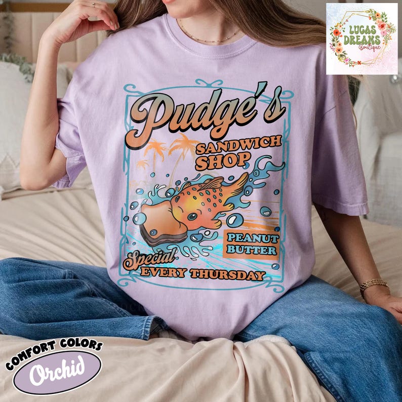 Pu&ograve; includere: T-shirt color orchidea con grafica retr&ograve; per "Pudge's Sandwich Shop". Il design presenta un sandwich di pesce a cartoni animati, palme e la scritta "Special Peanut Butter Every Thursday". La maglietta ha anche la scritta "Comfort Colors" e "Orchid".