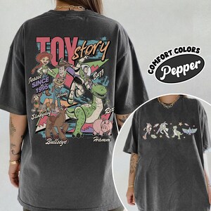 May include: Dark grey t-shirt with a vibrant Toy Story character graphic, including Woody, Buzz, and Rex. The back of the shirt features the words "Toy Story" and "Jessie Since 1995". The shirt also has the words "Comfort Colors Pepper" on the right side.