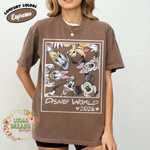 Op de afbeelding: Espresso-kleurig T-shirt met een Disney World-afbeelding met Mickey Mouse, Minnie Mouse, Donald Duck, Goofy en andere personages. Het shirt heeft de tekst "Disney World 2026" in een wit rechthoekig kader. Onderdeel van de "Comfort Colors" collectie.