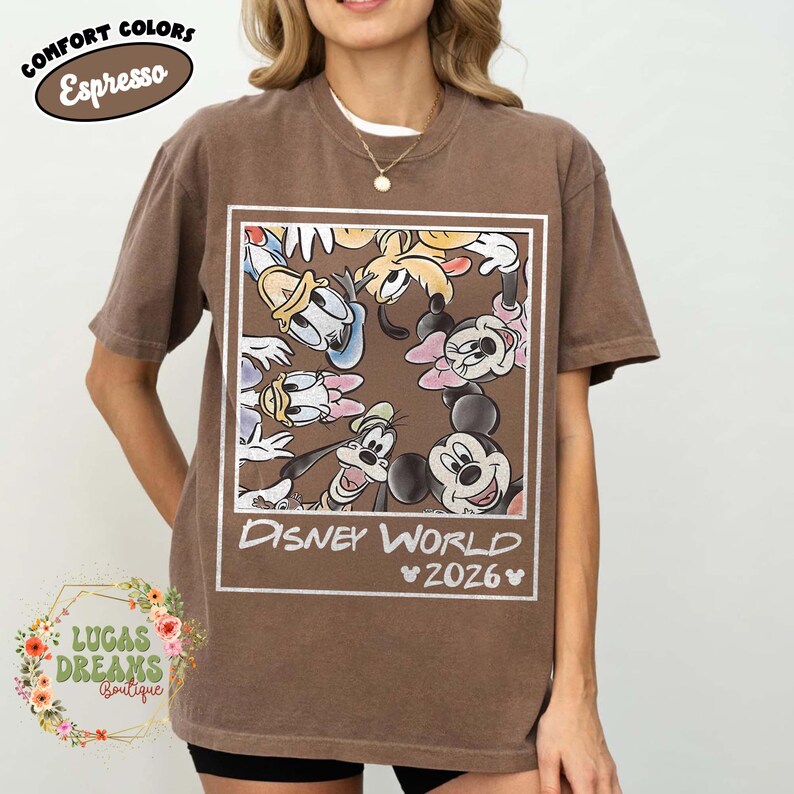 Op de afbeelding: Espresso-kleurig T-shirt met een Disney World-afbeelding met Mickey Mouse, Minnie Mouse, Donald Duck, Goofy en andere personages. Het shirt heeft de tekst "Disney World 2026" in een wit rechthoekig kader. Onderdeel van de "Comfort Colors" collectie.