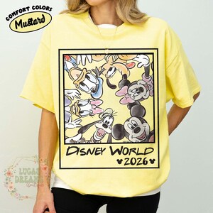Op de afbeelding: Mosterdgeel T-shirt met een ingelijste afbeelding van Disney-personages zoals Mickey Mouse, Minnie Mouse, Donald Duck en Goofy. De afbeelding luidt "Disney World 2026". Het shirt heeft korte mouwen en een ronde hals.