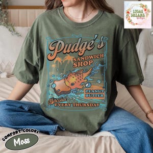 Pu&ograve; includere: T-shirt Comfort Colors verde muschio con grafica retr&ograve; per "Pudge's Sandwich Shop". Il design include un'illustrazione di sandwich al burro di arachidi, palme e il testo "Special Every Thursday."