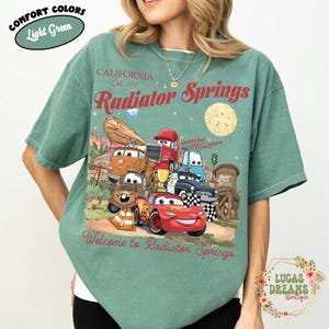 Pode incluir: Camiseta verde claro com um gr&aacute;fico de personagens do filme Carros, incluindo o Rel&acirc;mpago McQueen. A camiseta tem o texto "Radiator Springs" e "Welcome to Radiator Springs". A camiseta &eacute; da marca Comfort Colors.