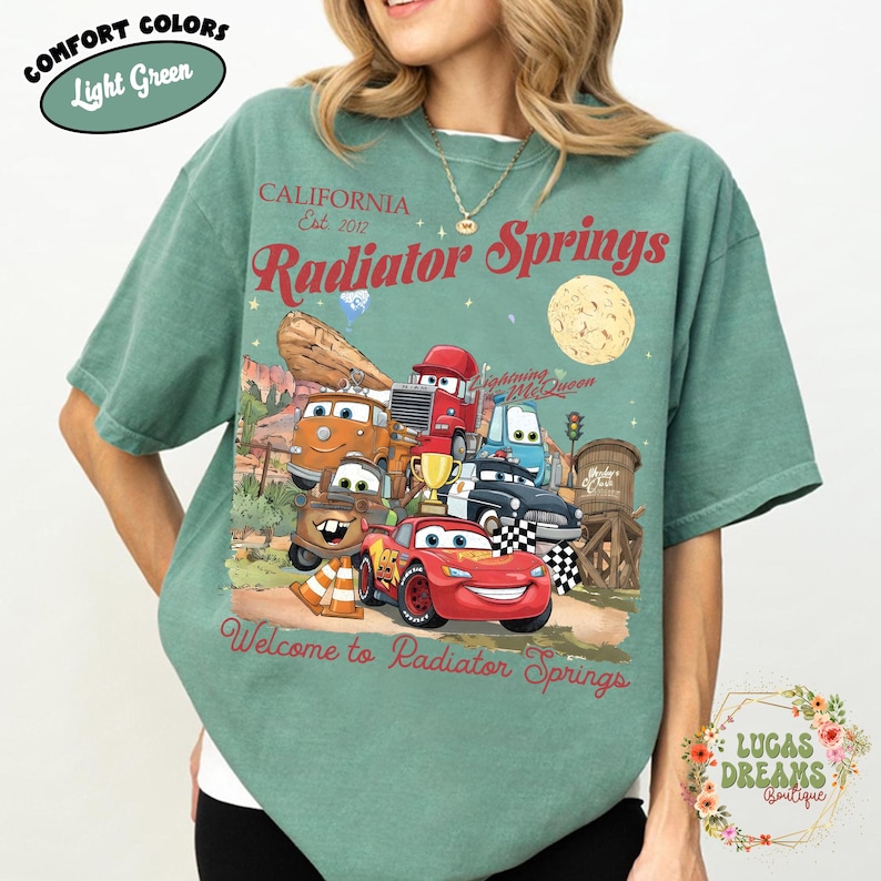 Pode incluir: Camiseta verde claro com um gr&aacute;fico de personagens do filme Carros, incluindo o Rel&acirc;mpago McQueen. A camiseta tem o texto "Radiator Springs" e "Welcome to Radiator Springs". A camiseta &eacute; da marca Comfort Colors.
