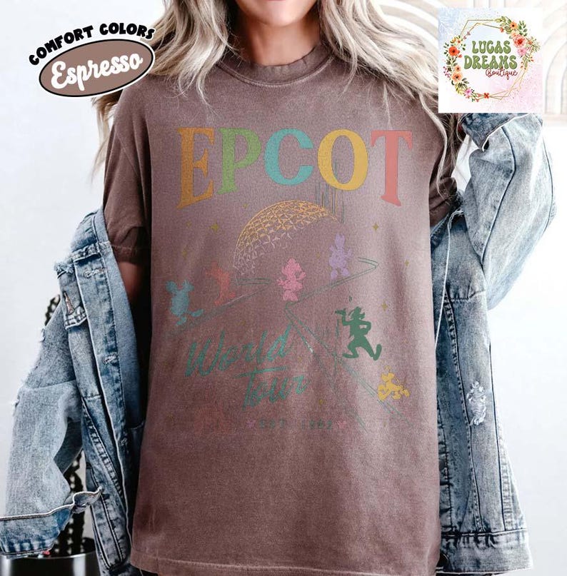 Puede incluir: Camiseta color espresso con la palabra "EPCOT" en letras coloridas. La camiseta presenta un gr&aacute;fico de la esfera EPCOT y personajes de dibujos animados. Las palabras "World Tour" est&aacute;n impresas debajo. La camiseta es parte de la colecci&oacute;n Comfort Colors.