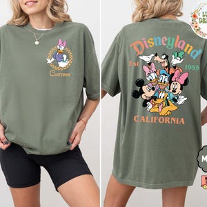 Puede incluir: Camiseta verde musgo con Daisy Duck en la parte delantera. La parte trasera de la camiseta muestra un colorido dise&ntilde;o de Disneyland con Mickey, Minnie, Donald y Goofy. Tambi&eacute;n se ve el texto "Disneyland California".